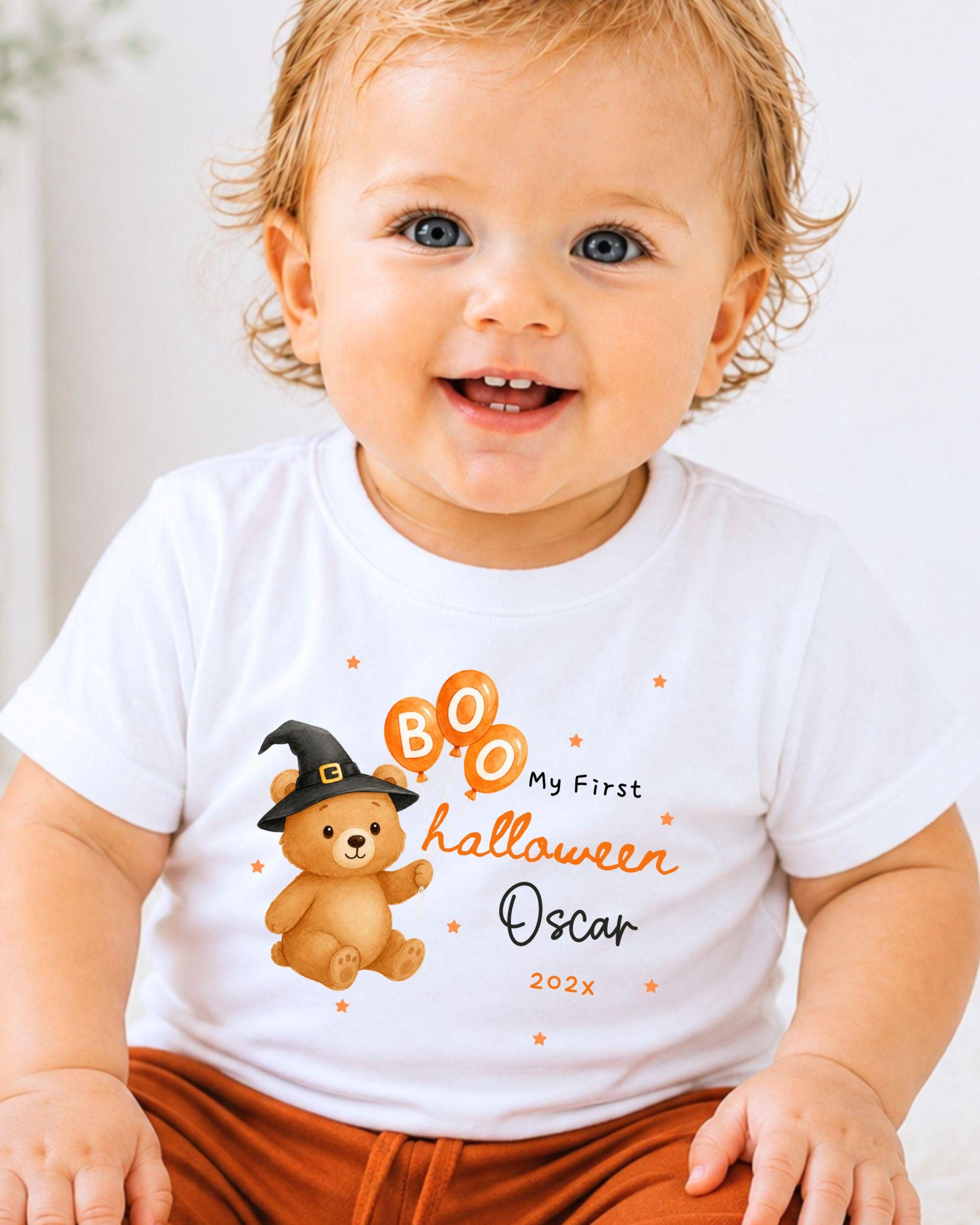 A smiling baby wearing a white t-shirt with a Halloween-themed design and text.