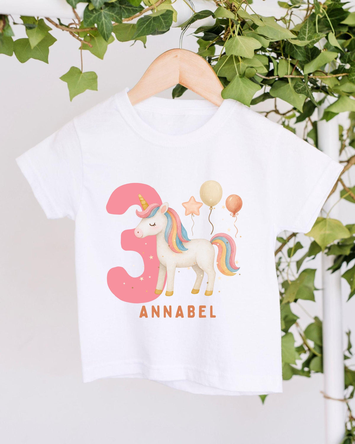 A white t-shirt with a unicorn and balloons printed on it, hanging on a wooden hanger.