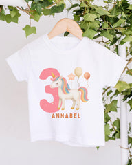 A white t-shirt with a unicorn and balloons printed on it, hanging on a wooden hanger.