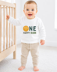 A smiling baby boy wearing a white sweatshirt with a smiley face and the text "ONE HAPPY Dude" is standing next to a wooden crib.