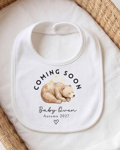 Personalisable white Bib with a sleeping brown watercolour bear beneath curved black 'Coming Soon' lettering, 'Baby Owen Autumn 2027' text and an outlined heart.