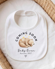 A white bib with a printed design of a sleeping bear and the text "COMING SOON Baby Owen Autumn 2027" printed on it.