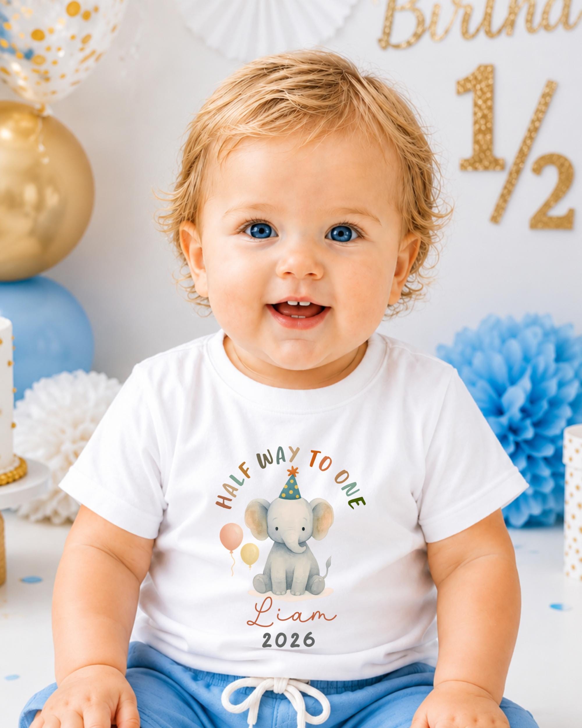 A smiling baby boy wearing a white t-shirt with a graphic of an elephant and the text "Half Way to One" and "Liam" printed on it, sitting in front of a birthday party decoration.