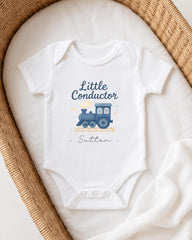 A white baby onesie with a blue train graphic and the text "Little Conductor Sutton" printed on it, placed on a white blanket inside a woven basket.
