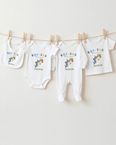 A row of white baby clothes with the text "One-DER the sea" printed on them, along with the name "Joshua" printed below.