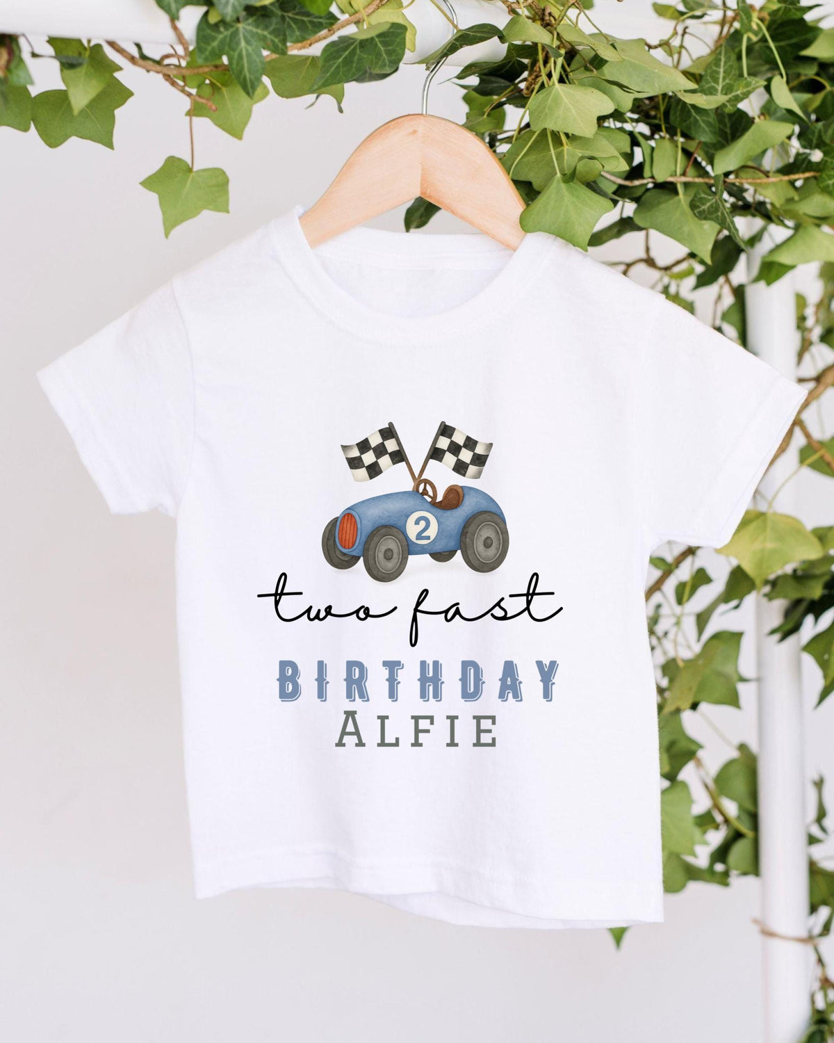 A white t-shirt with a graphic of a race car and the text "two fast birthday alfie" printed on it, hanging on a wooden hanger.