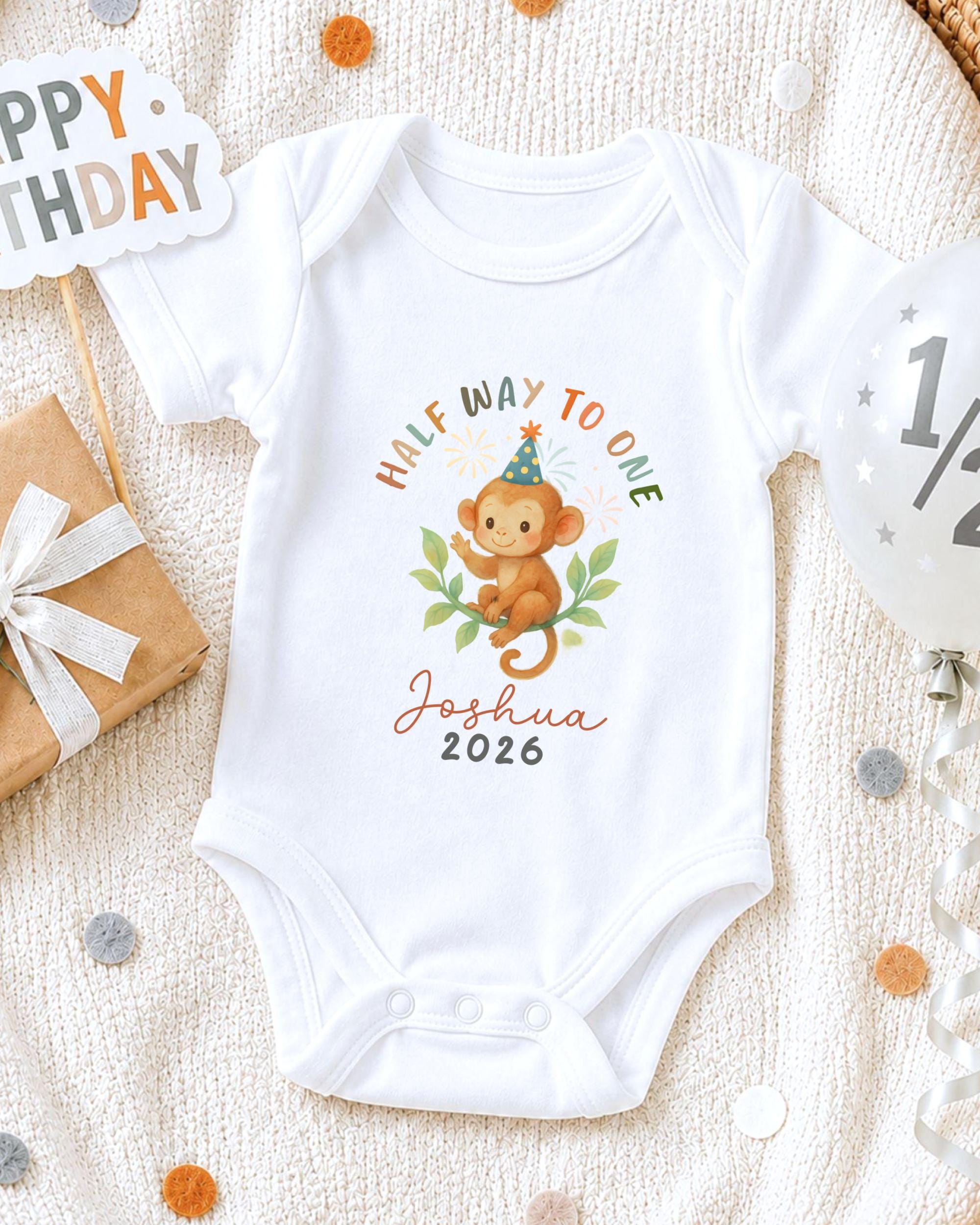 A white onesie with a monkey graphic and the text "Half Way to One" printed on it, along with the name "Joshua" and the year "2026". The onesie is placed on a white blanket, surrounded by birthday decorations such as a "Happy Birthday" sign, a balloon, and confetti.