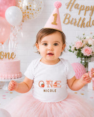 A young girl wearing a pink birthday hat and a white t-shirt with the name "Nicole" is posing for a birthday photo. The background includes balloons, a cake, and flowers, creating a festive atmosphere.