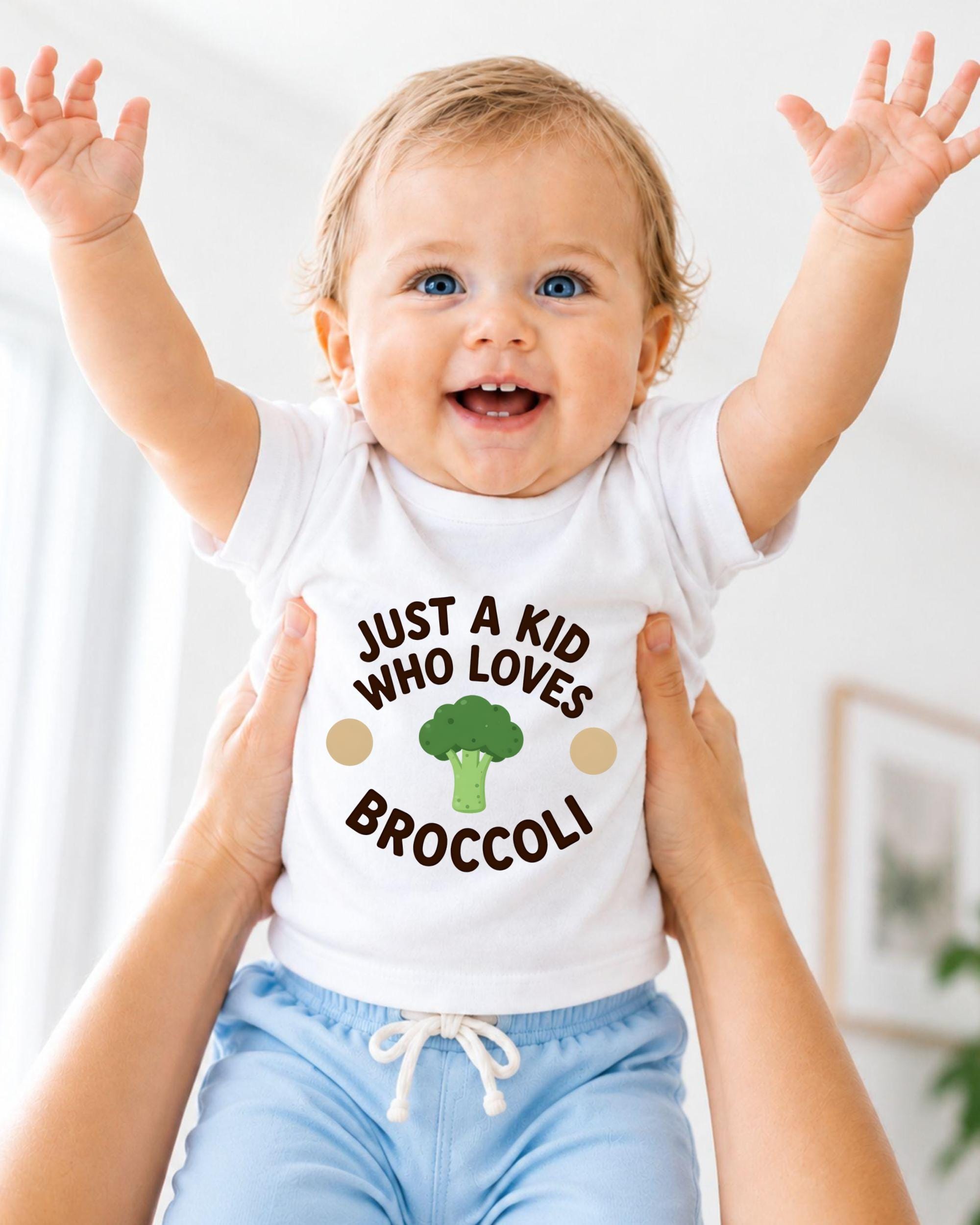 A smiling baby wearing a white shirt with the text "Just a kid who loves broccoli" is being held up by someone's arms.