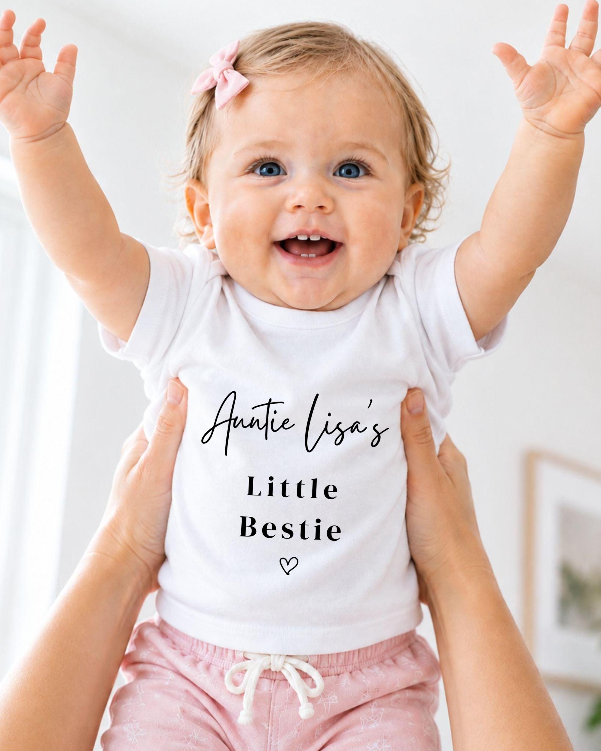 A smiling baby girl wearing a white t-shirt with the text "Auntie Lisa's Little Bestie" and pink shorts, being held up by someone's arms.