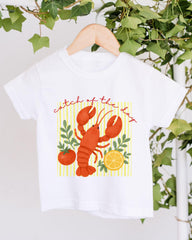 A white t-shirt with a graphic design featuring a lobster and various fruits, including a lemon and a tomato, along with the text "Catch of the day".