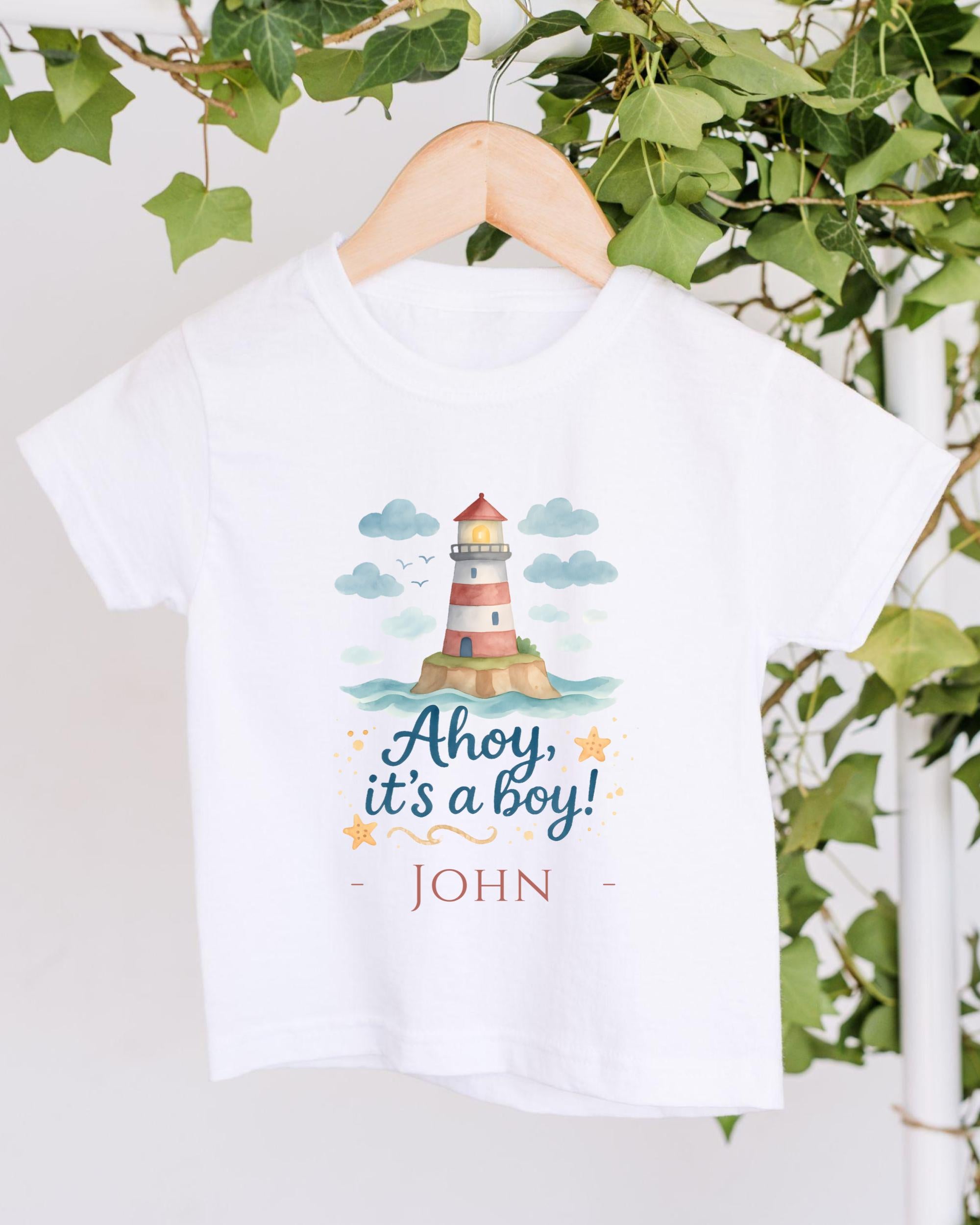 A white t-shirt with a lighthouse graphic and the text "Ahoy, it's a boy!" printed on it, hanging on a wooden hanger.