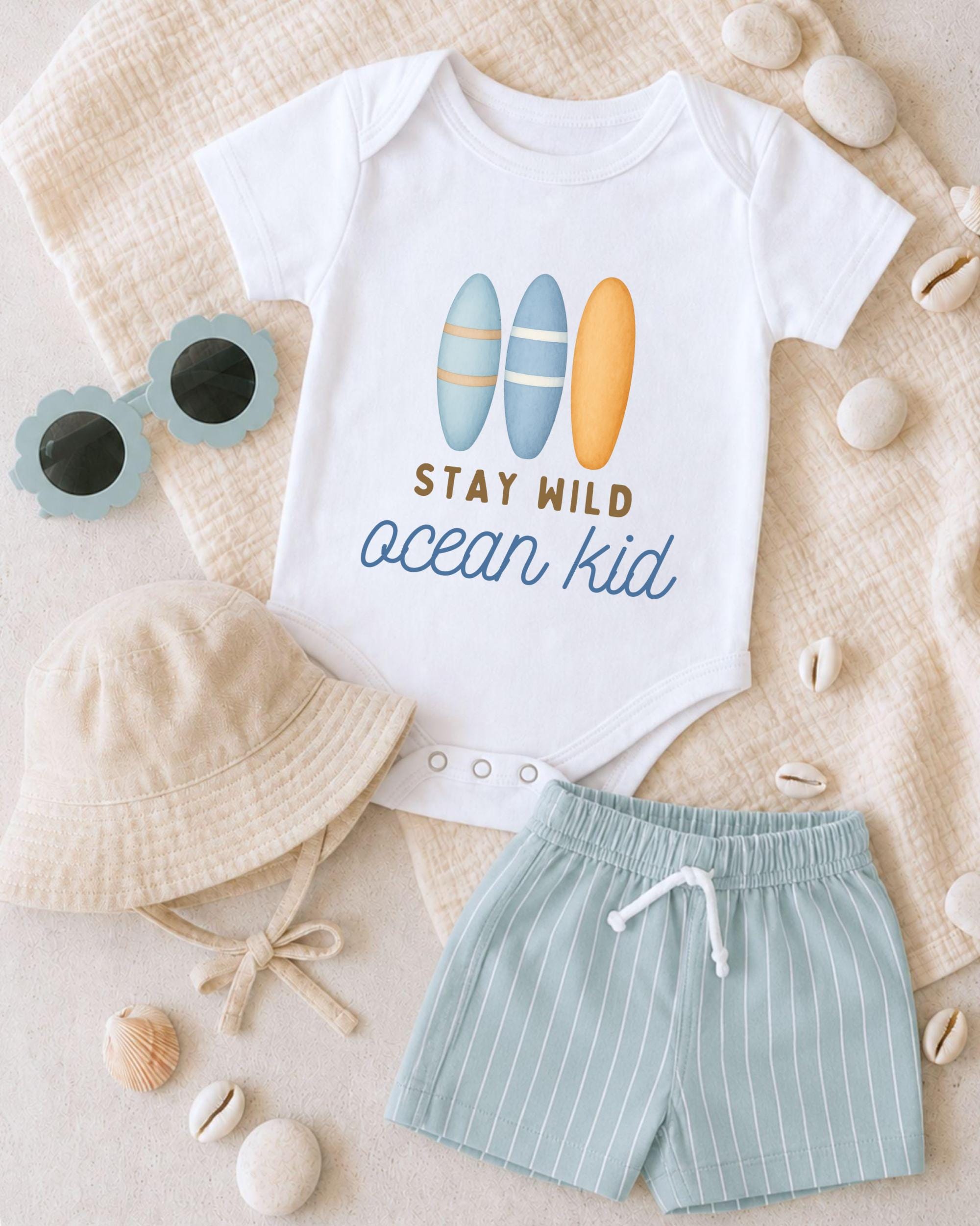 A baby's outfit, including a white onesie with a surfboard design, a beige bucket hat, and striped shorts, along with sunglasses and seashells, is laid out on a beige surface.