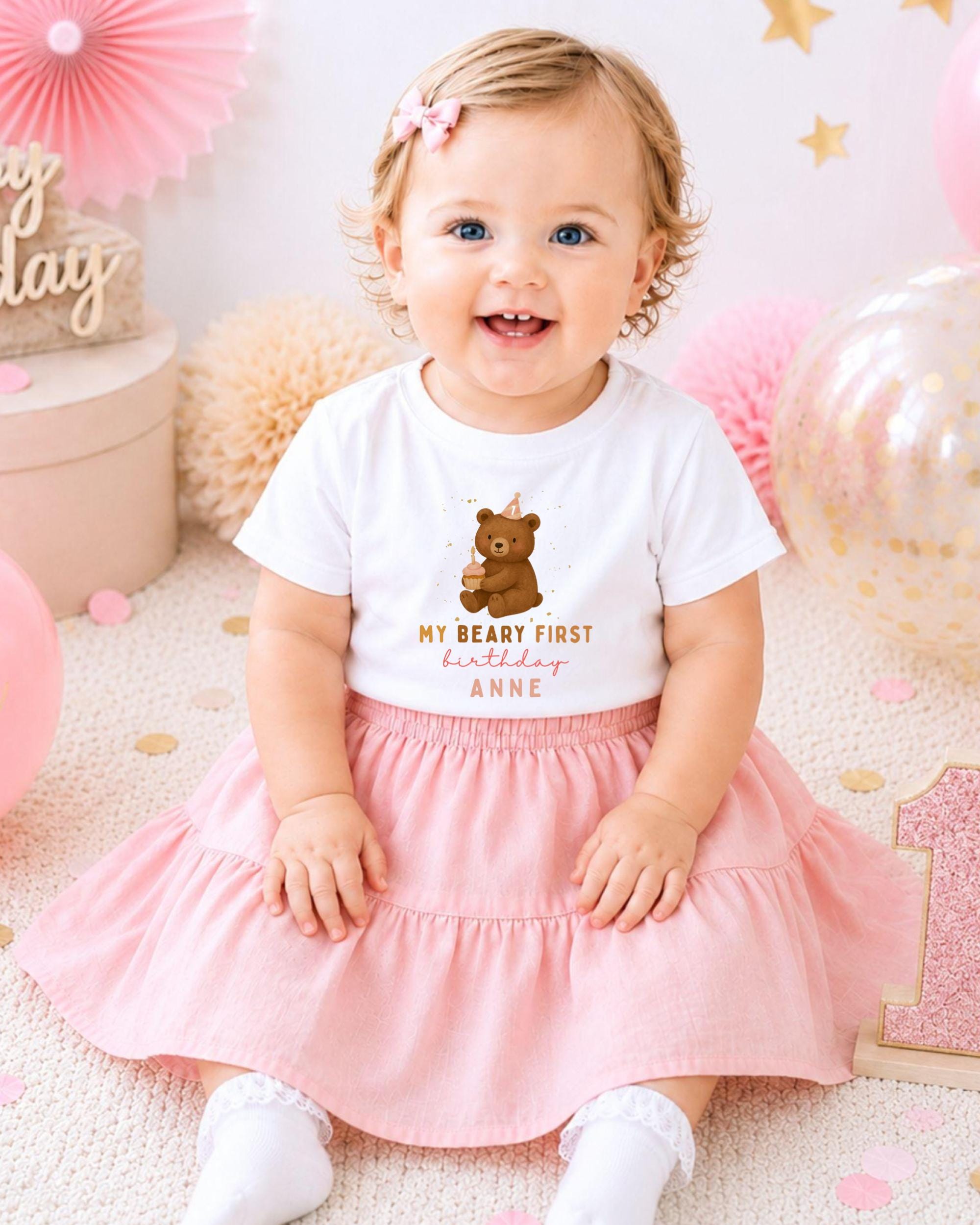 A smiling baby girl wearing a white t-shirt with a teddy bear graphic and the text "My Beary First Birthday Anne" sits on a pink and white background with balloons and confetti.