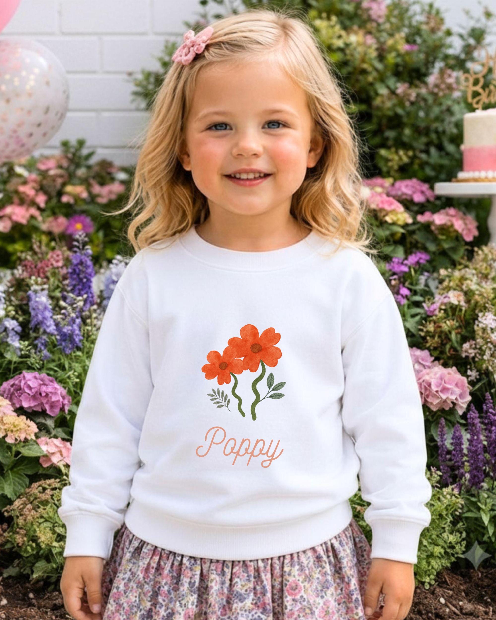 A young girl with blonde hair is smiling and wearing a white sweater with a flower design and the word "Poppy" printed on it. She is standing in front of a garden filled with colorful flowers.