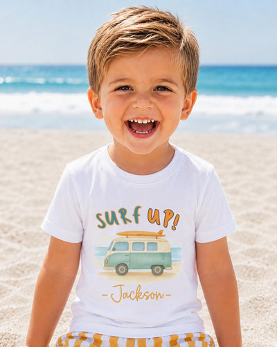 A young boy with a big smile on his face, wearing a white t-shirt with a surfboard graphic and the text "Surf up! Jackson" printed on it, standing on a sandy beach with the ocean in the background.