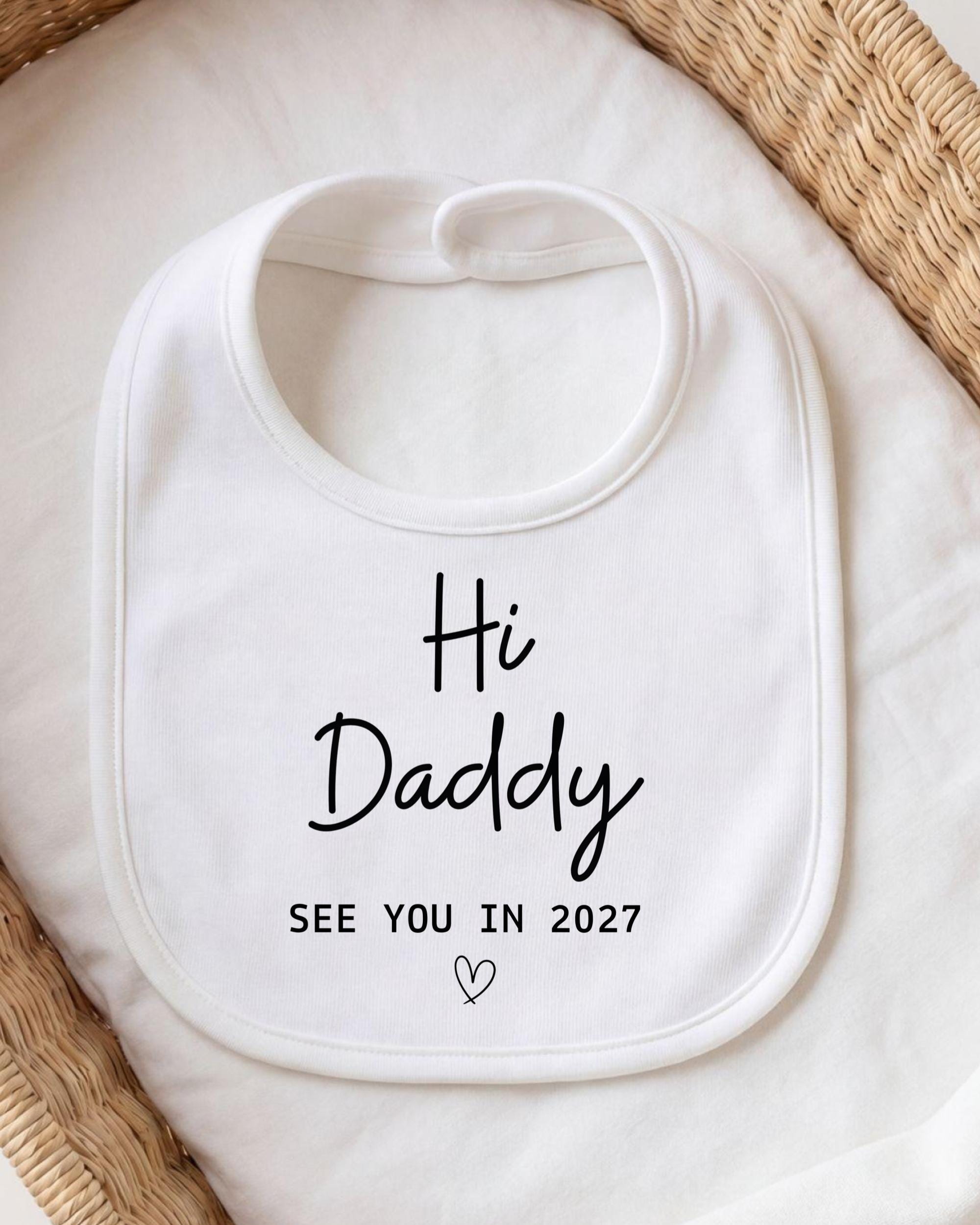 A white bib with the text "Hi Daddy See You in 2027" printed on it, placed on a white cloth.