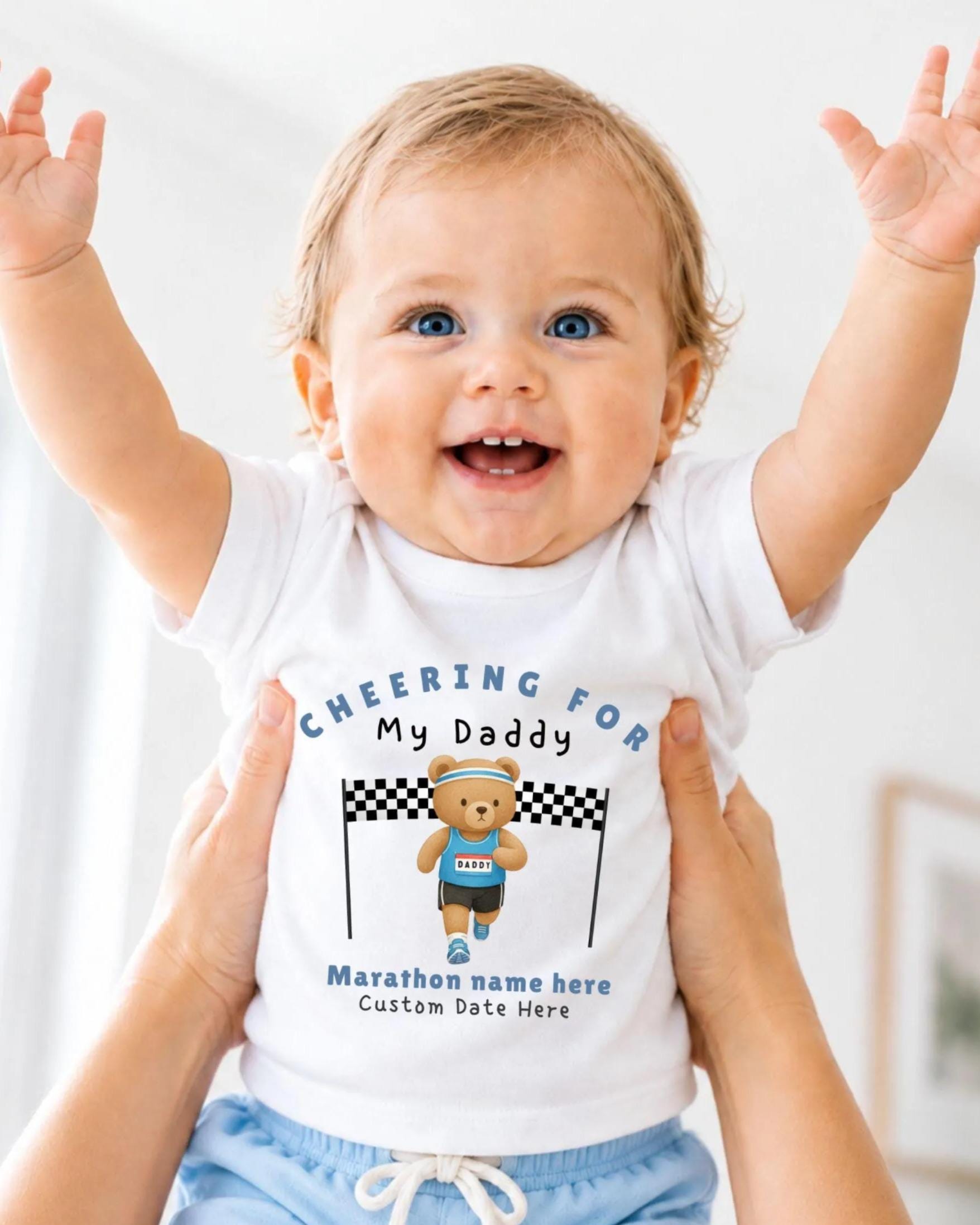 A smiling baby wearing a white t-shirt with a teddy bear and text, being held up by someone's arms.
