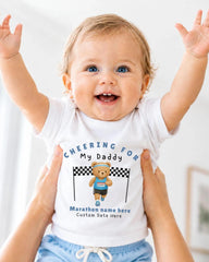 A smiling baby wearing a white t-shirt with a teddy bear and text, being held up by someone's arms.