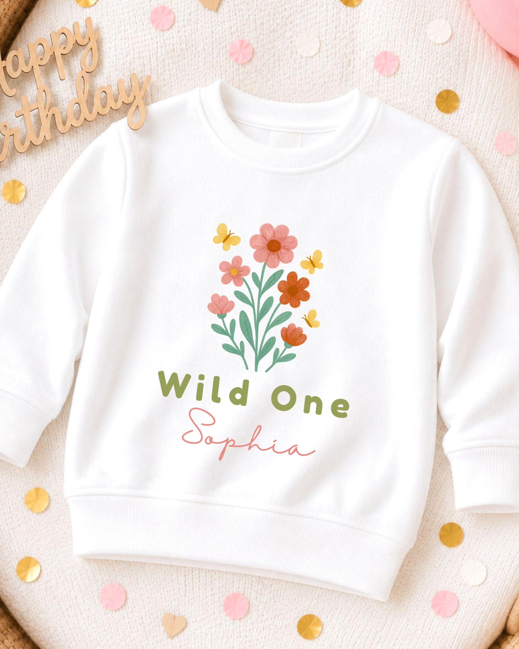 A white sweatshirt with a floral design and the text "Wild One Sophia" printed on it, surrounded by confetti.