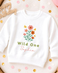 A white sweatshirt with a floral design and the text "Wild One Sophia" printed on it, surrounded by confetti.