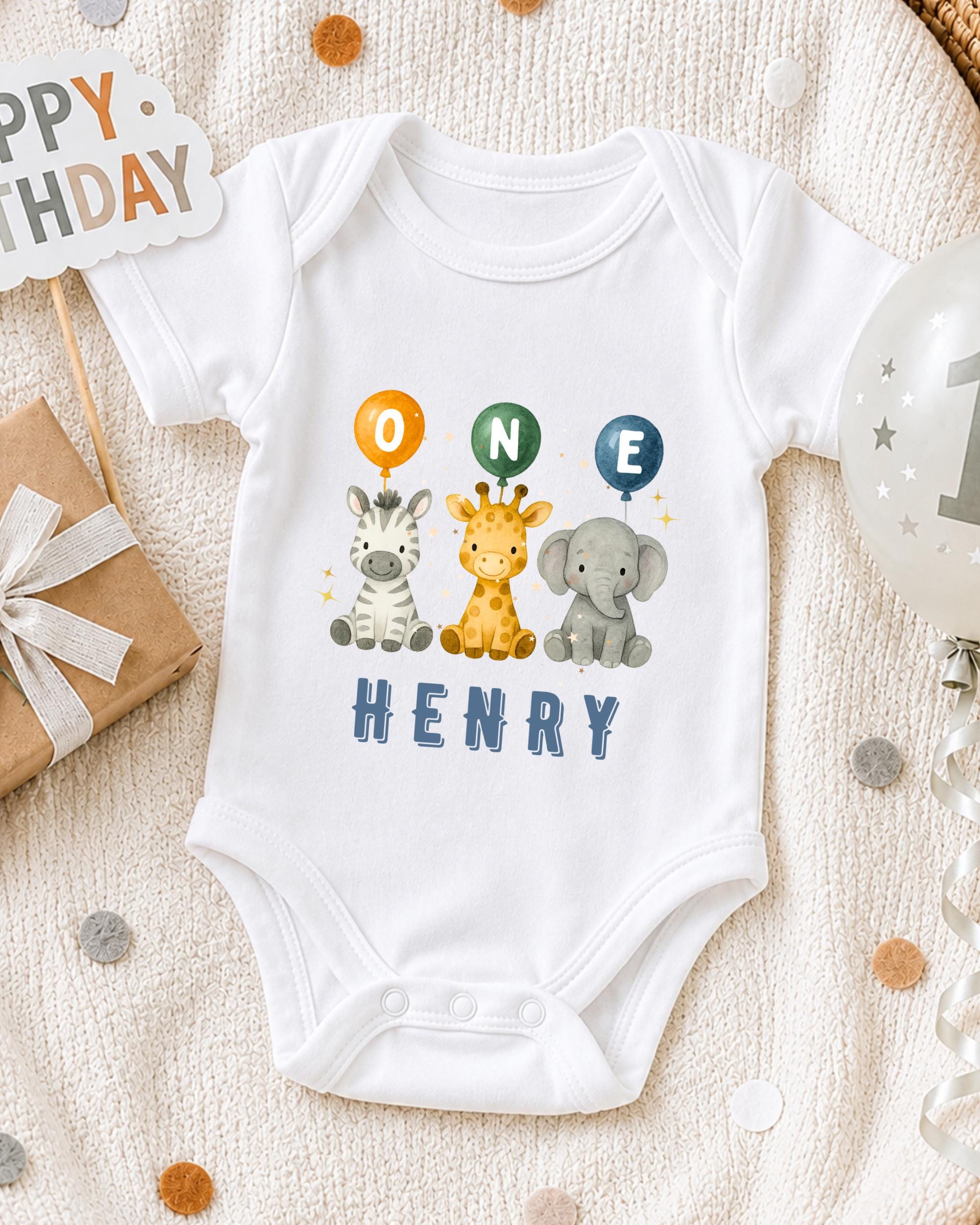 A white onesie with a cartoon image of a giraffe, zebra, and elephant, along with the text "ONE HENRY" printed on it, is displayed on a bed with a "Happy Birthday" sign and a gift box.