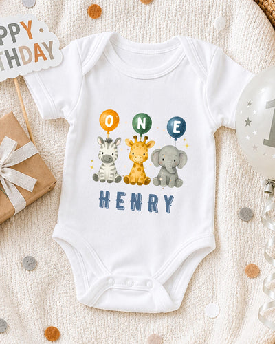 A white bodysuit with a cartoon image of a giraffe, zebra, and elephant, along with the text "ONE HENRY" printed on it, is displayed on a bed with a "Happy Birthday" sign and a gift box.