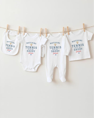 A row of four white baby clothes with the text "WATCHING TENNIS DADDY Jackson" printed on them, hanging on a clothesline.