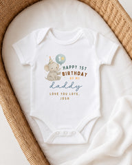 A white onesie with a cartoon elephant and text that reads "Happy 1st Birthday as my Daddy. Love You Lots. Josh" is displayed on a white blanket.