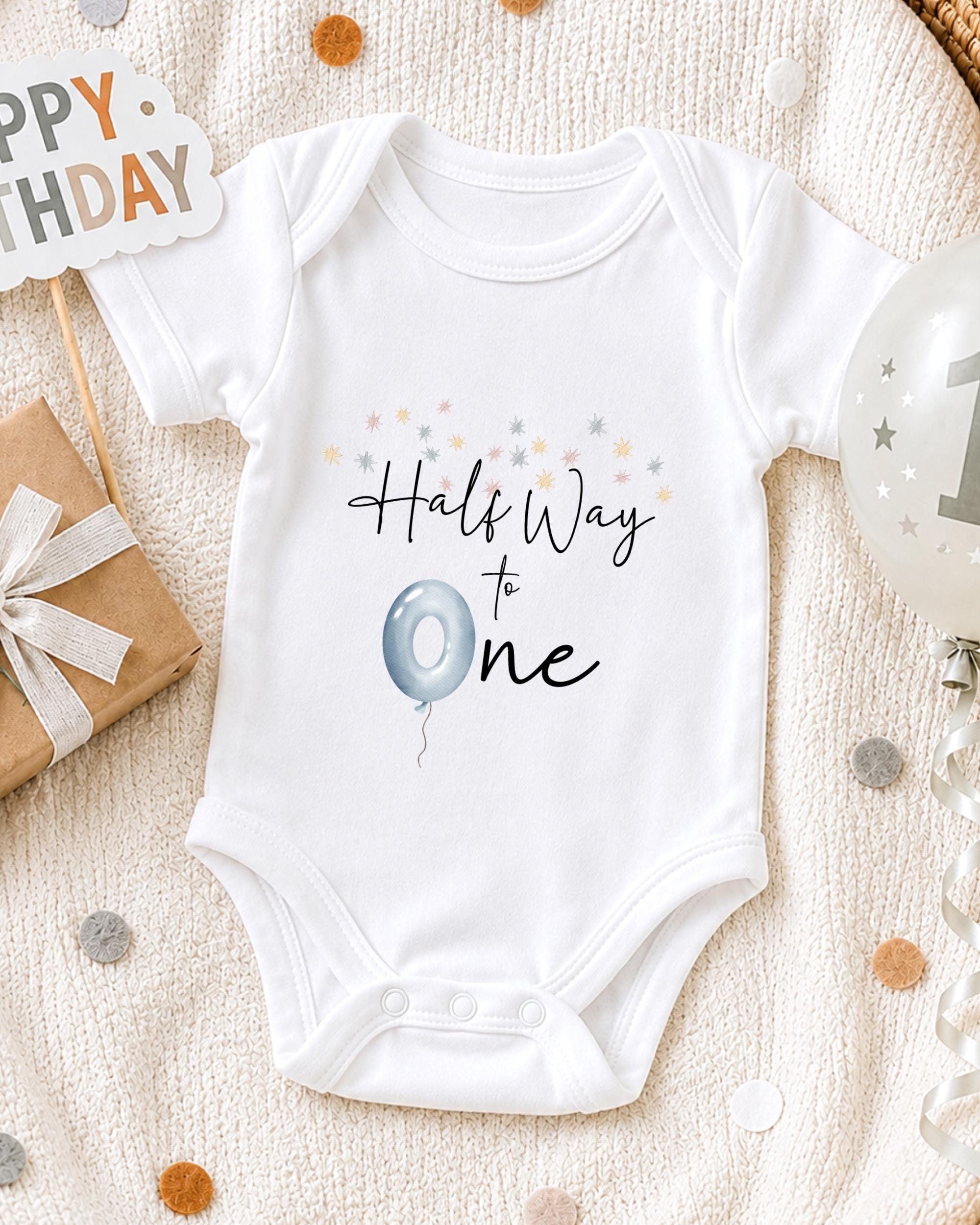 A white onesie with the text "Half Way to One" printed on it, along with a blue balloon and confetti, is displayed on a bed.