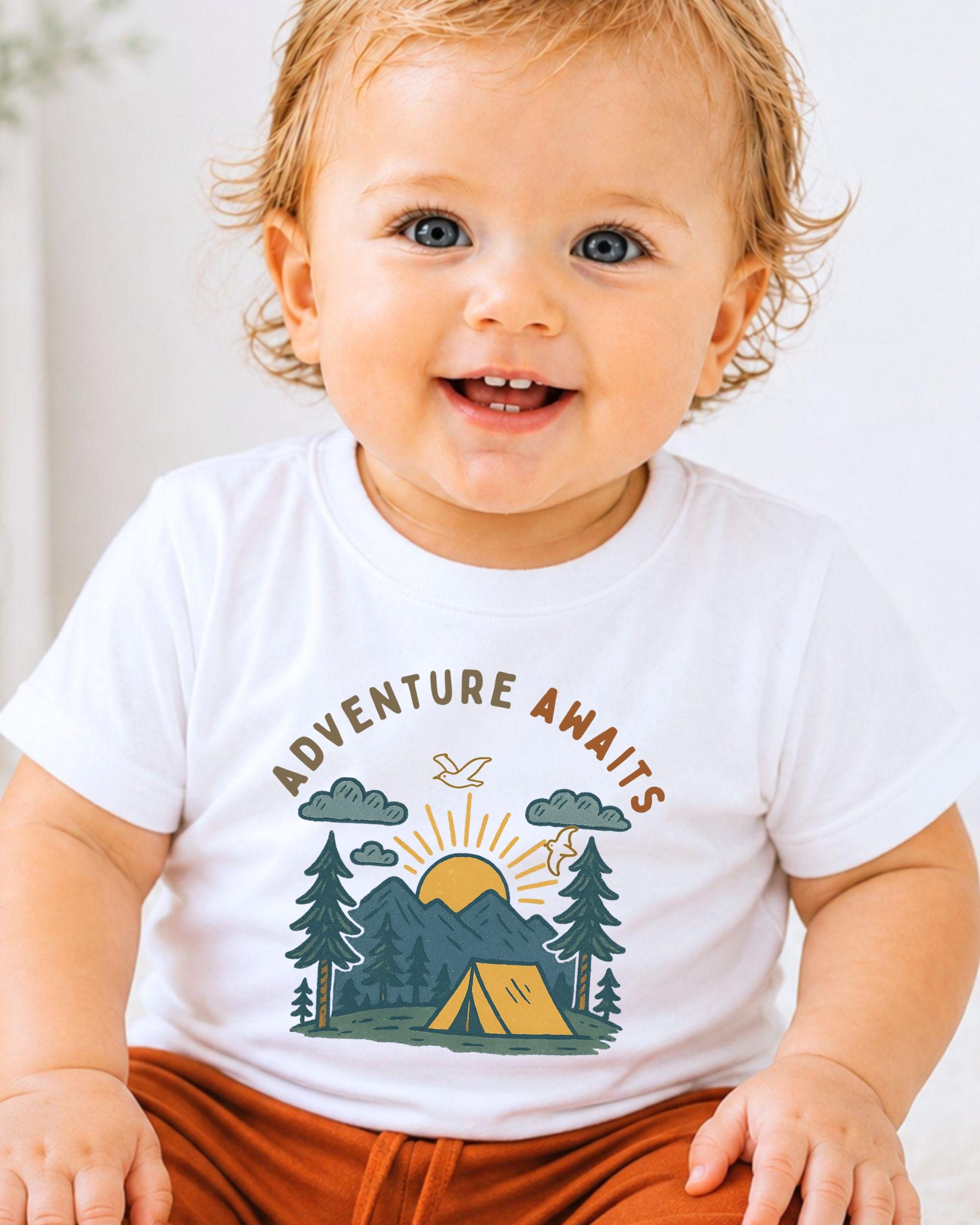 A smiling baby wearing a white t-shirt with a graphic design of a camping scene and the text "Adventure Awaits".
