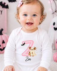 A smiling baby girl wearing a white onesie with a pink bat and a ghost character on it, sitting in front of Halloween decorations.