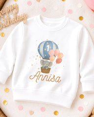 A white sweatshirt with a cartoon elephant and a hot air balloon design, along with the name 'Annisa' printed on it.