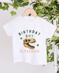 A white t-shirt with a dinosaur skull and the text "BIRTHDAY BOY ALEXANDER" printed on it, hanging on a wooden hanger.