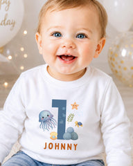 A smiling baby boy wearing a white shirt with a personalized number 1 and ocean-themed design.
