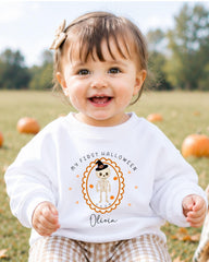A smiling baby girl wearing a white sweater with a Halloween-themed design and text.