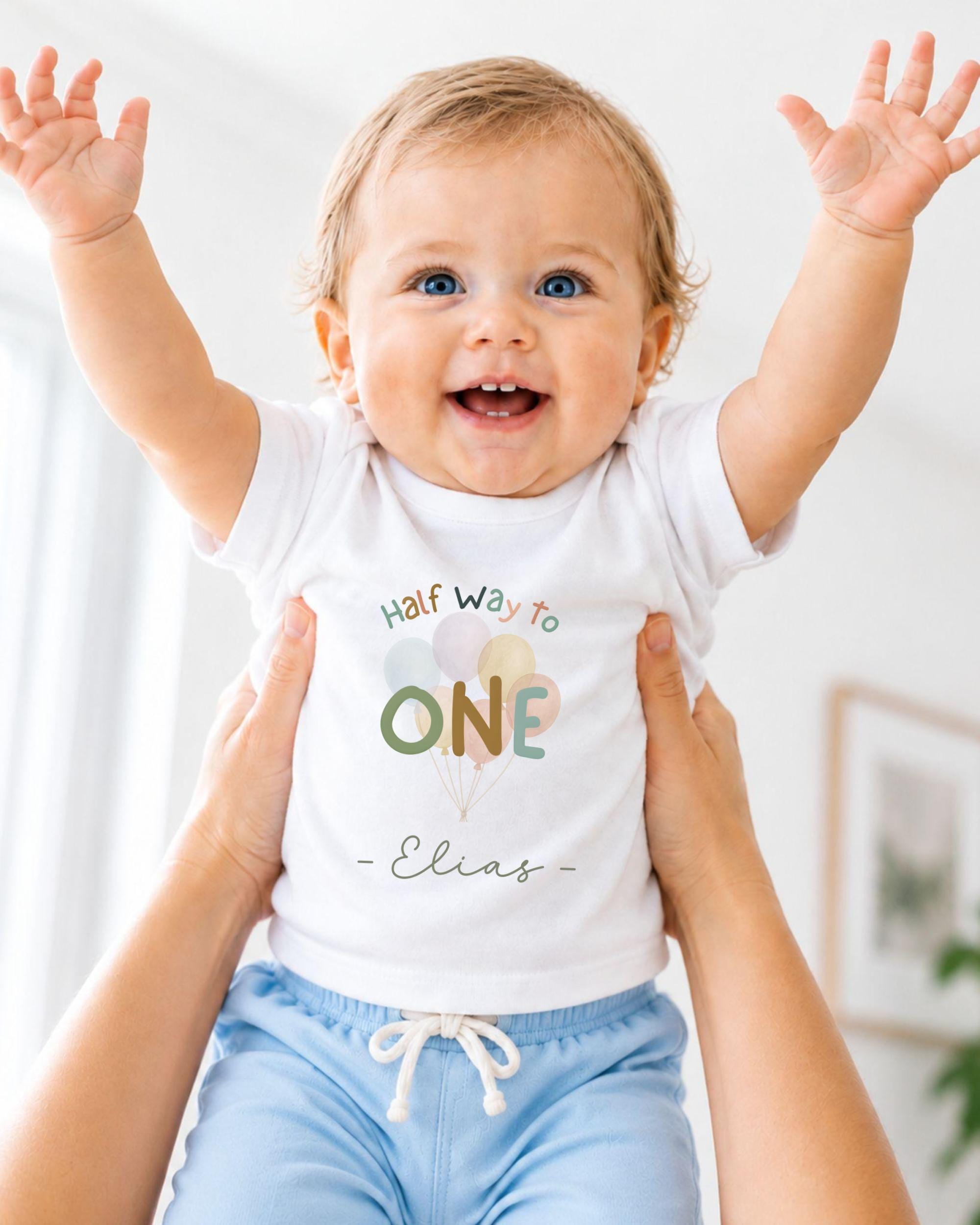 A smiling baby wearing a white t-shirt with the text "Half Way to One - Elias" is being held up by someone's arms.