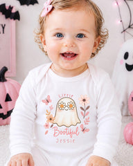 A smiling baby girl wearing a white onesie with a ghost and floral design, sitting in front of Halloween decorations.