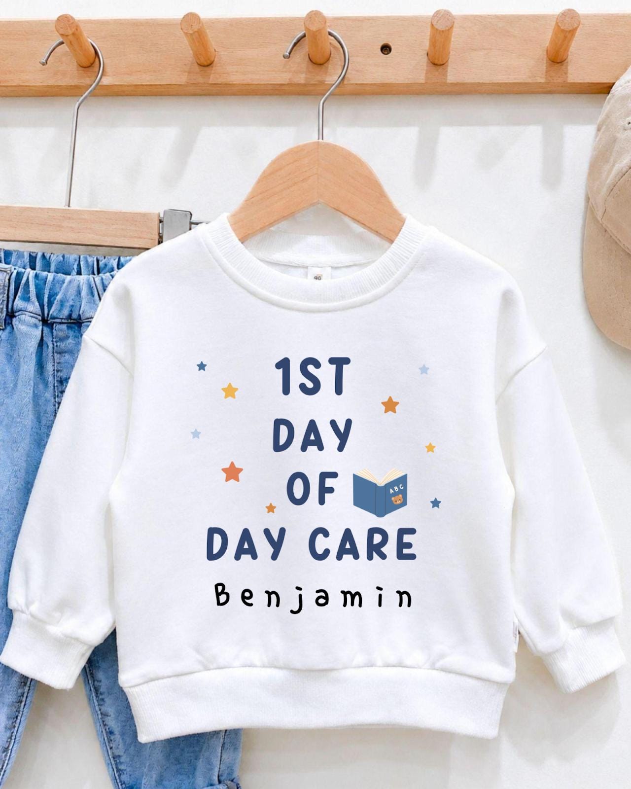 A white sweatshirt with the text "1st DAY OF DAY CARE" printed on it, along with a small illustration of a book and stars.