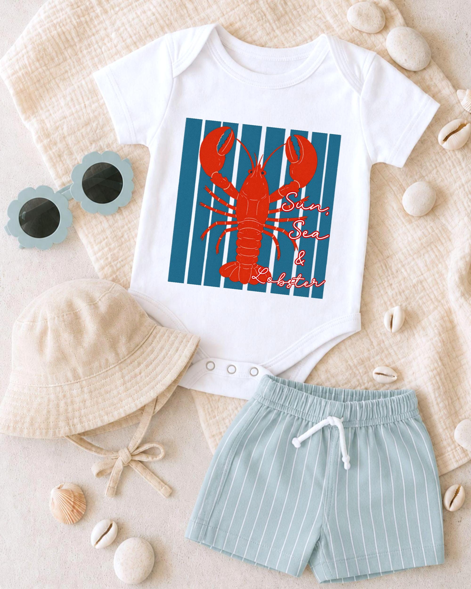 A white baby onesie with a red lobster design, a beige bucket hat, and a pair of light blue shorts, along with some sunglasses and seashells, are arranged on a beige surface.