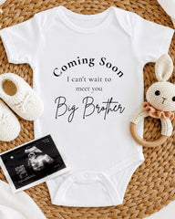 A white onesie with text that says "Coming Soon I can't wait to meet you Big Brother" is displayed on a woven surface, accompanied by a crocheted bunny toy and a photograph of an Ultrasound image.