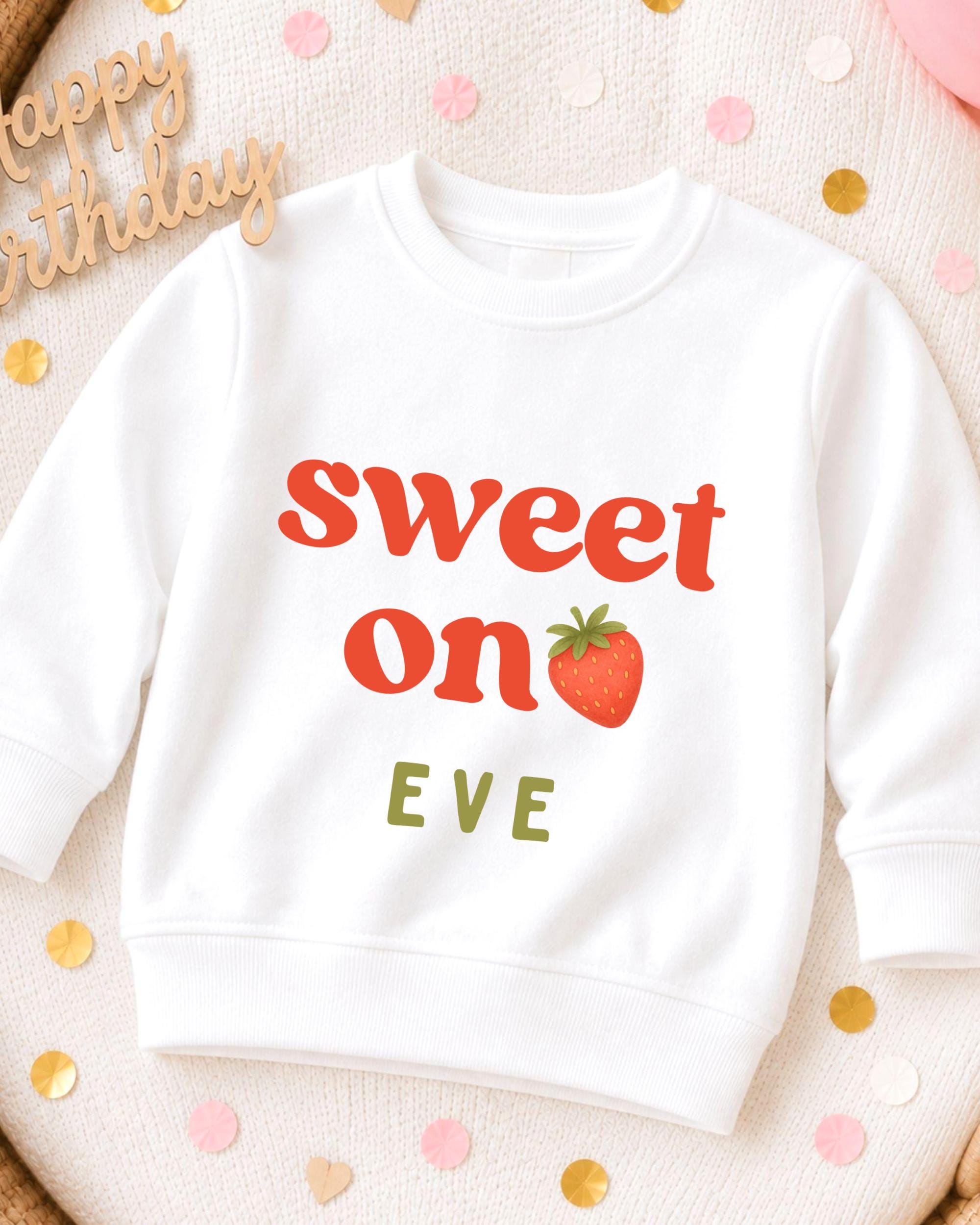 A white sweatshirt with the text "Sweet on Eve" printed on it, along with a strawberry graphic. The sweatshirt is placed on a surface with scattered confetti.