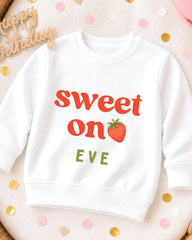A white sweatshirt with the text "Sweet on Eve" printed on it, along with a strawberry graphic. The sweatshirt is placed on a surface with scattered confetti.