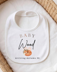 A white baby bib with the text "BABY Wood Arriving Autumn 26" printed on it, along with an illustration of a sleeping fox.