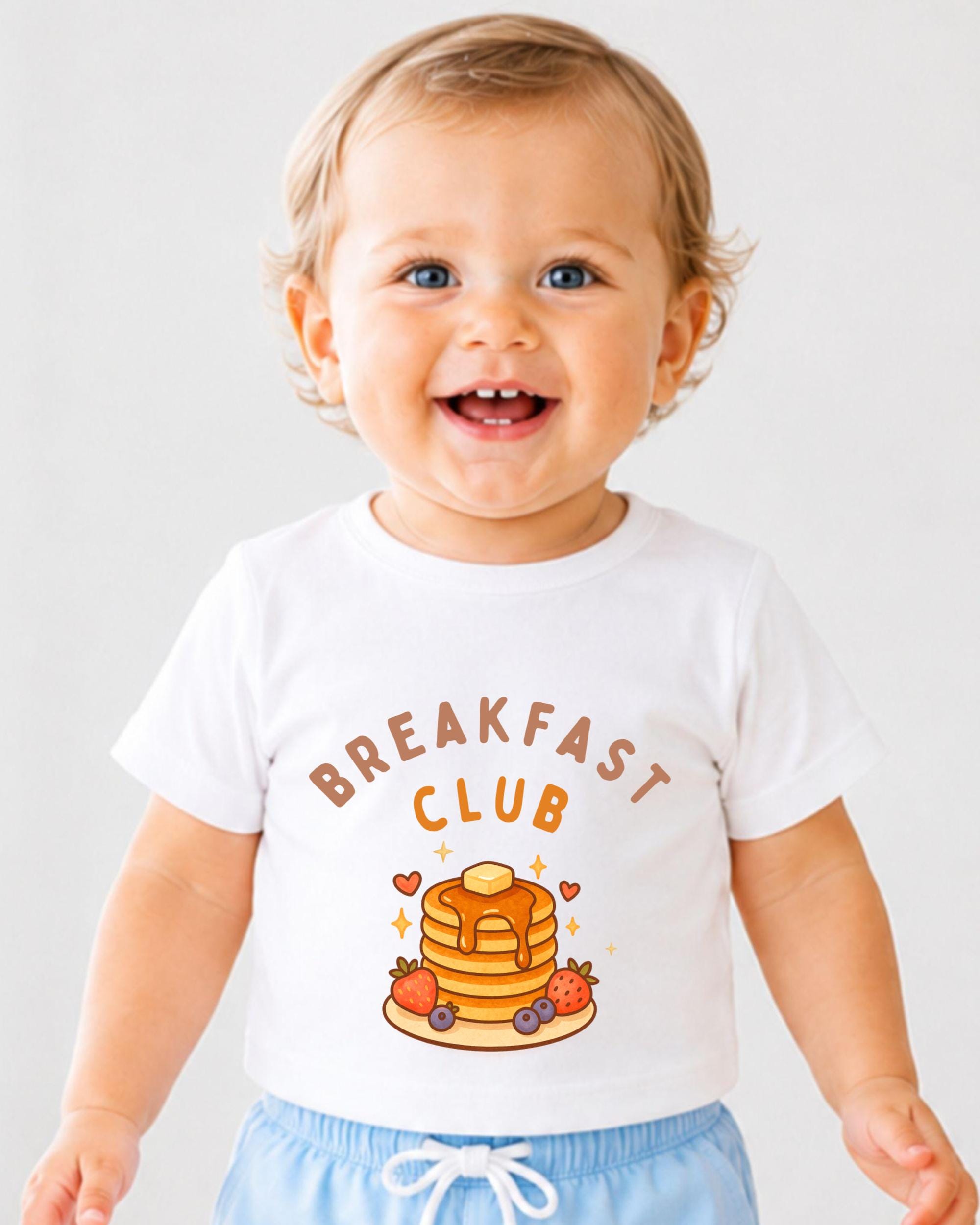 A smiling baby wearing a white t-shirt with a breakfast club graphic and text.