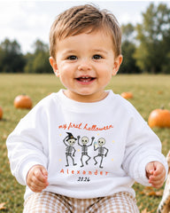 A smiling baby boy wearing a white sweatshirt with a Halloween-themed design and text.