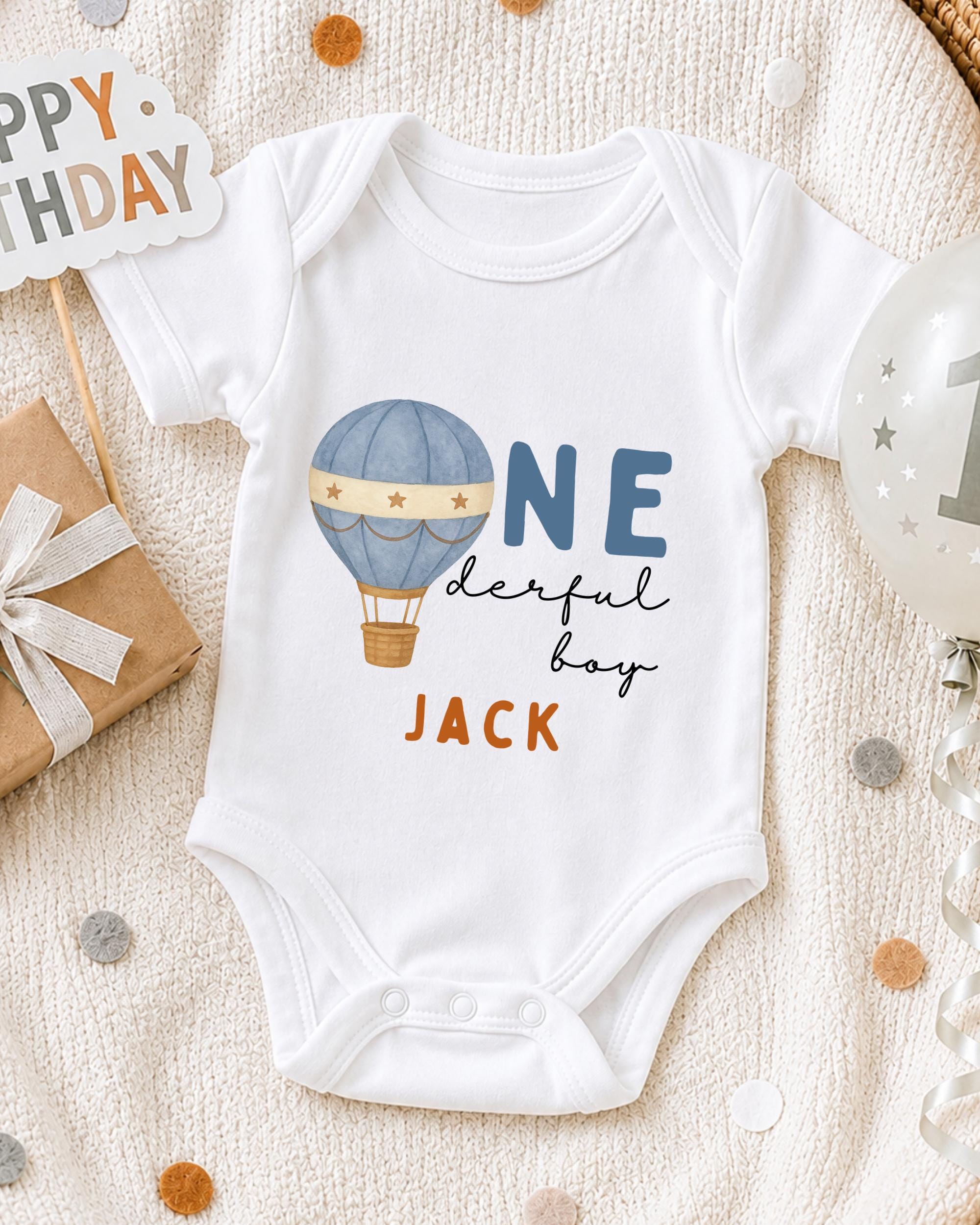A white onesie with a hot air balloon design and the text "ONE derful boy" printed on it, along with a birthday sign and confetti.