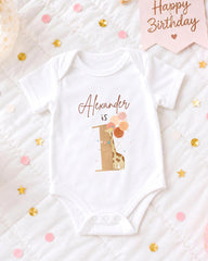 A white onesie with a giraffe design and the text "Alexander is" printed on it, placed on a bed with confetti and a birthday card.
