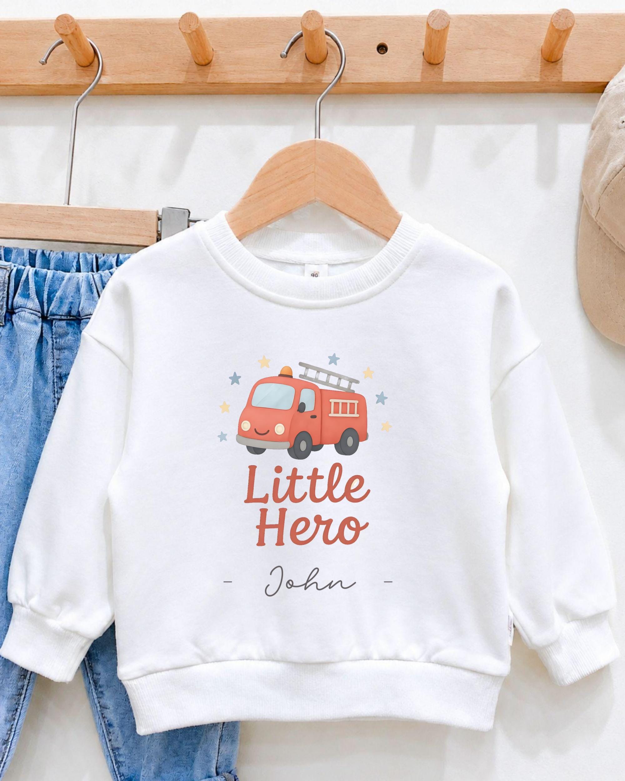A white sweatshirt with a red fire truck graphic and the text "Little Hero John" printed on it, hanging on a wooden hanger.