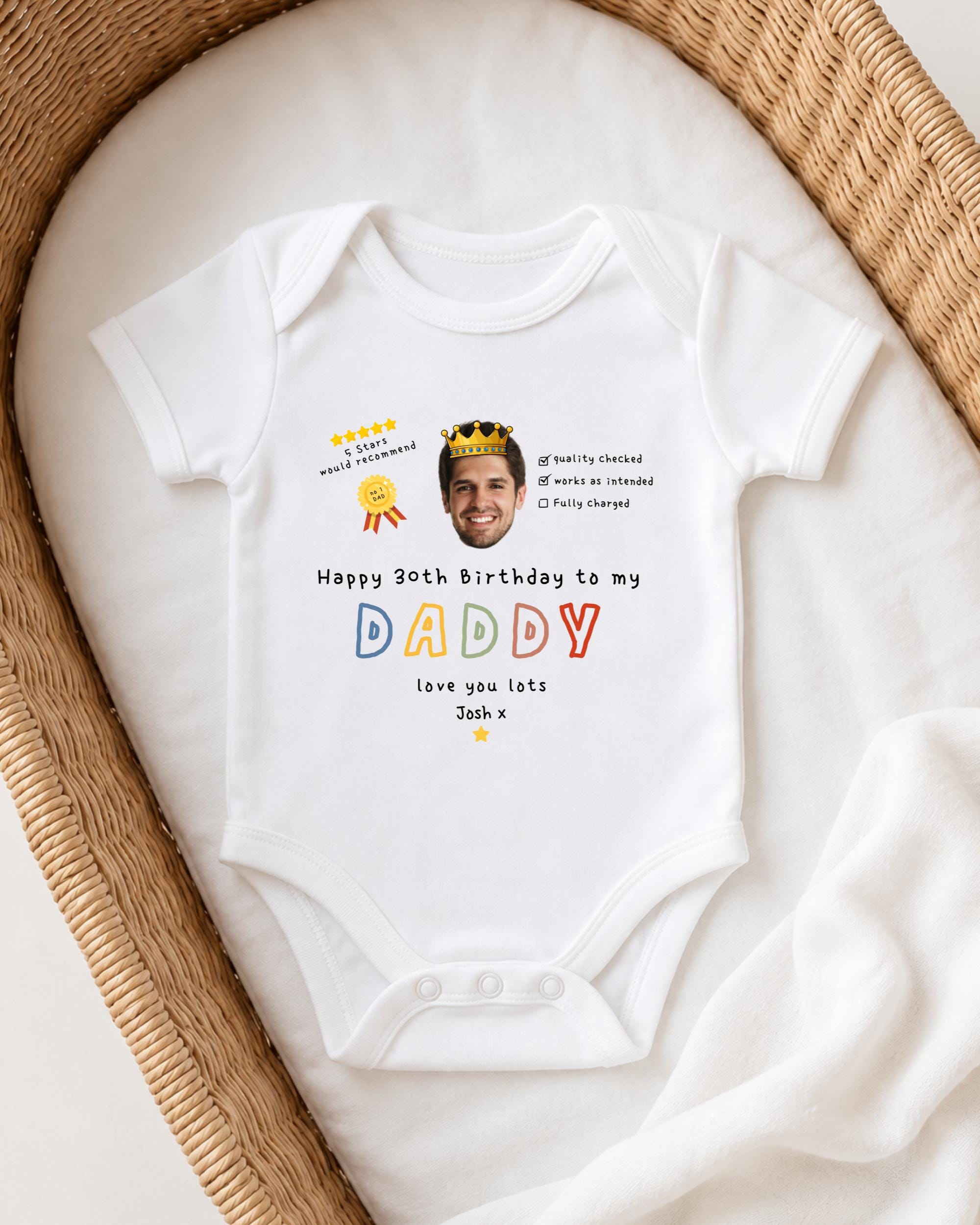 A white baby onesie with a personalized message and a crown on it, placed on a white blanket.